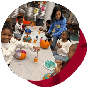 A group of students paint pumpkins with a teacher while sitting at a table.