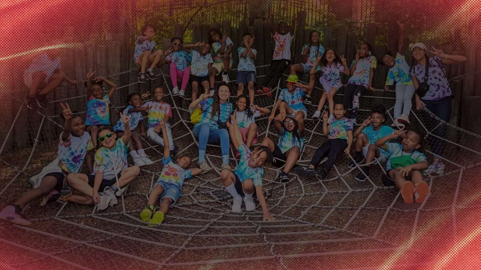 A large group of students and two teachers pose on a giant web at the zoo.